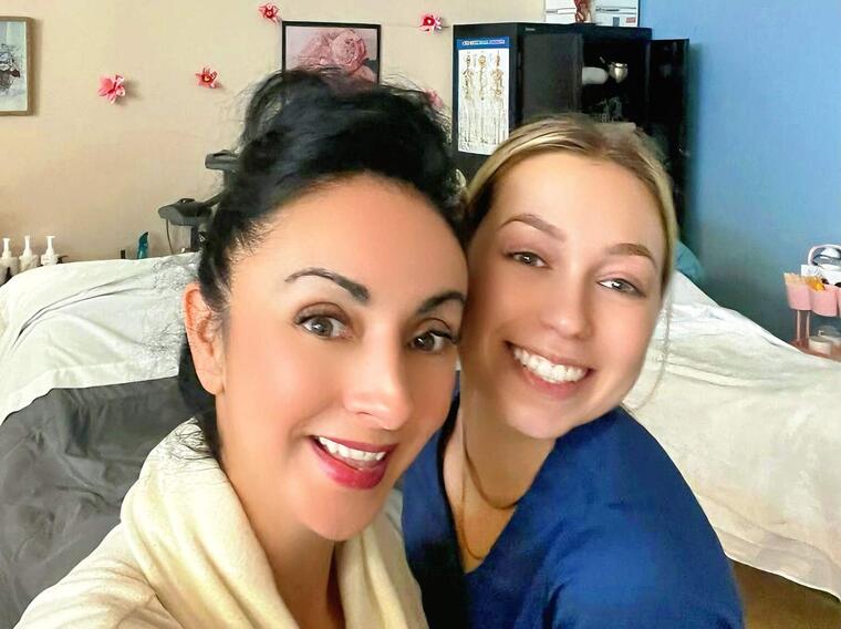 Two women smile in a medical esthetics classroom filled with spa beds.