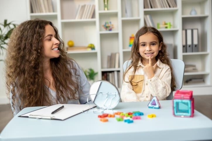 A female early childcare assistant helping a smiling female pupil with her schoolwork